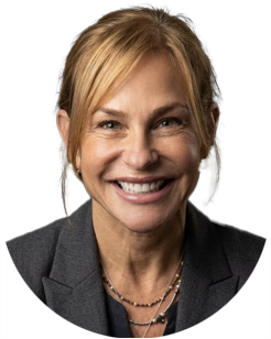 Smiling woman with light brown hair in a low ponytail, wearing a gray blazer and layered necklaces, against a plain white background.