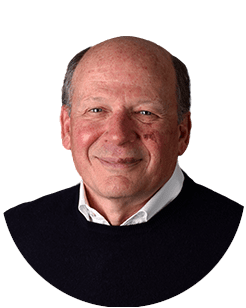 Balding older man with a friendly smile, wearing a white shirt and black sweater, against a white background.