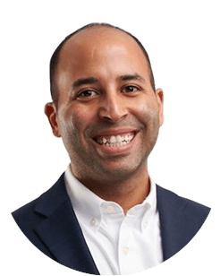A man in a dark suit and white shirt is smiling against a white background.