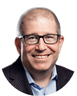 A bald man with glasses smiles, wearing a dark jacket over a light blue shirt, isolated on a white background.