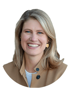 A person with shoulder-length blonde hair, wearing a tan coat with black buttons and a white shirt, is smiling.