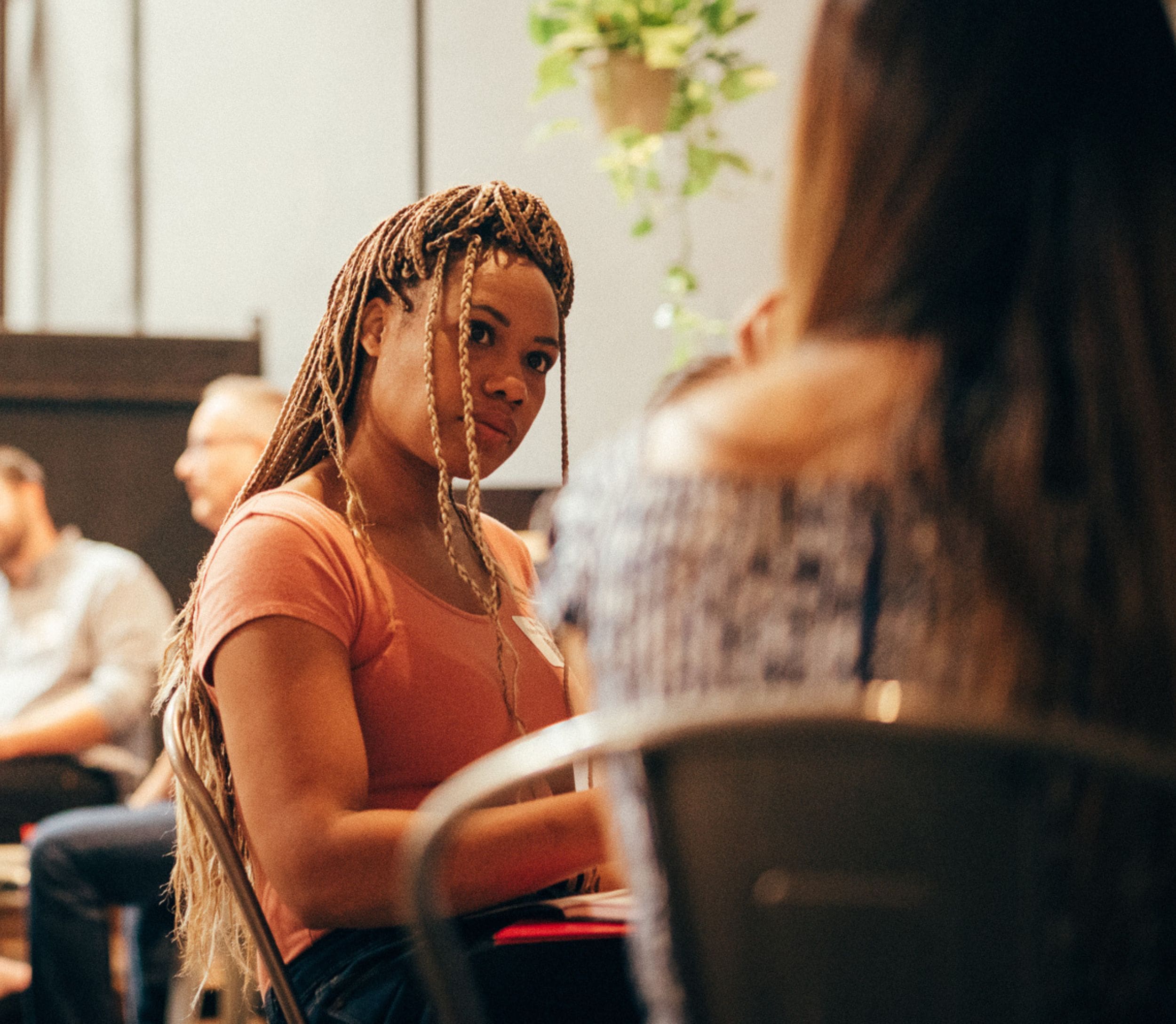 A woman with long braids in a pink top sits indoors, facing another person, with others sitting in the background.