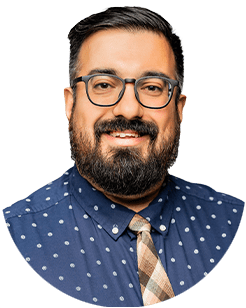 A man with a beard and glasses, wearing a navy blue polka dot shirt and patterned tie, smiles at the camera against a white background.