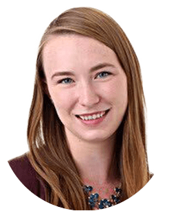 A woman with long, straight hair and a slight smile wearing a blue necklace and a brown top is pictured in front of a plain white background.