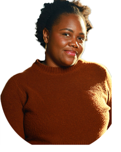 A person with short curly hair wearing a brown sweater stands against a plain light background, looking at the camera with a slight smile.