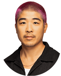 A person with short, dyed pink hair and a closely trimmed beard looks directly at the camera, wearing a black collared shirt and a gold necklace.