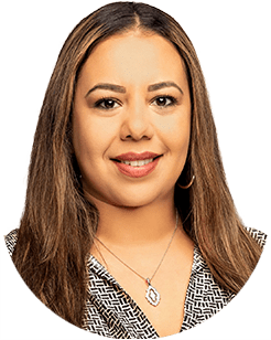A woman with long, straight hair and wearing a patterned blouse with a necklace smiles at the camera in a headshot.