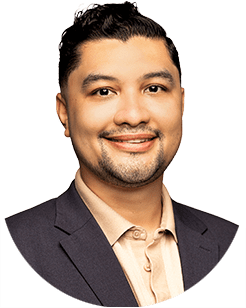 A man with dark hair and a trimmed beard is smiling, wearing a light patterned shirt and a dark blazer. The background is plain white.