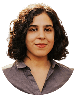 A person with shoulder-length curly hair and a neutral expression, wearing a gray collared shirt.