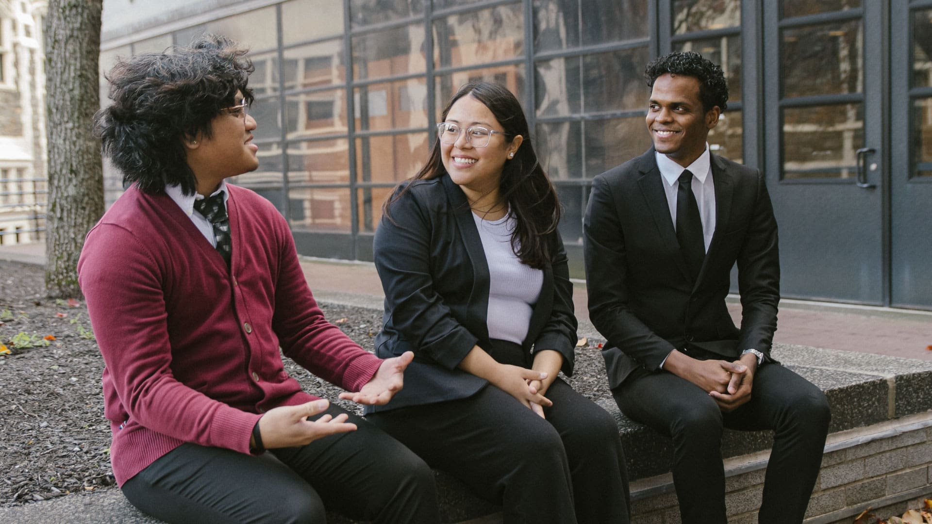 Three young adults sitting and chatting outdoors, one in a red cardigan and bowtie, one in glasses and a black sweater, and one in a black suit, all smiling.