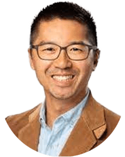 A person with short black hair and glasses is smiling, wearing a light blue shirt and a tan blazer.