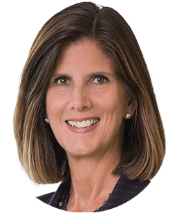 A woman with shoulder-length hair and brown highlights, smiling and wearing a striped blazer and pearl earrings.