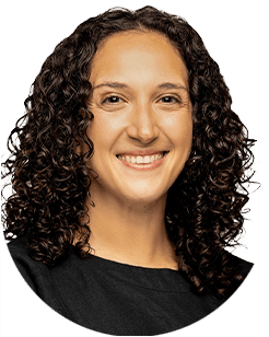 A person with curly hair smiles at the camera, wearing a black top, against a plain white background.