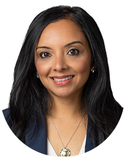 A woman with long black hair wearing a dark blazer and a necklace smiles in a headshot.