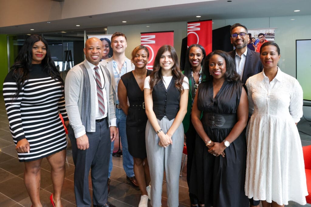 A group of nine people, dressed in business casual and formal attire, stand indoors posing for a photo. They are positioned in two rows with a conference-style backdrop.