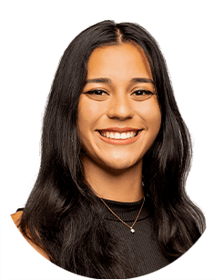 Headshot of a smiling individual with long, dark hair, wearing a black top and a necklace with a small pendant, against a plain white background.