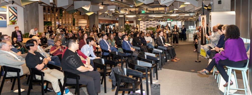 A group of people seated in rows listen attentively to a panel of speakers during a presentation in a modern conference room.