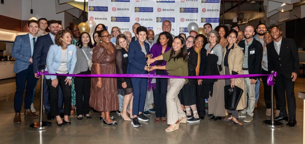 A group of people stand together smiling at a ribbon-cutting ceremony. A woman in the foreground cuts the ribbon with oversized scissors. A banner in the background displays logos of sponsors.