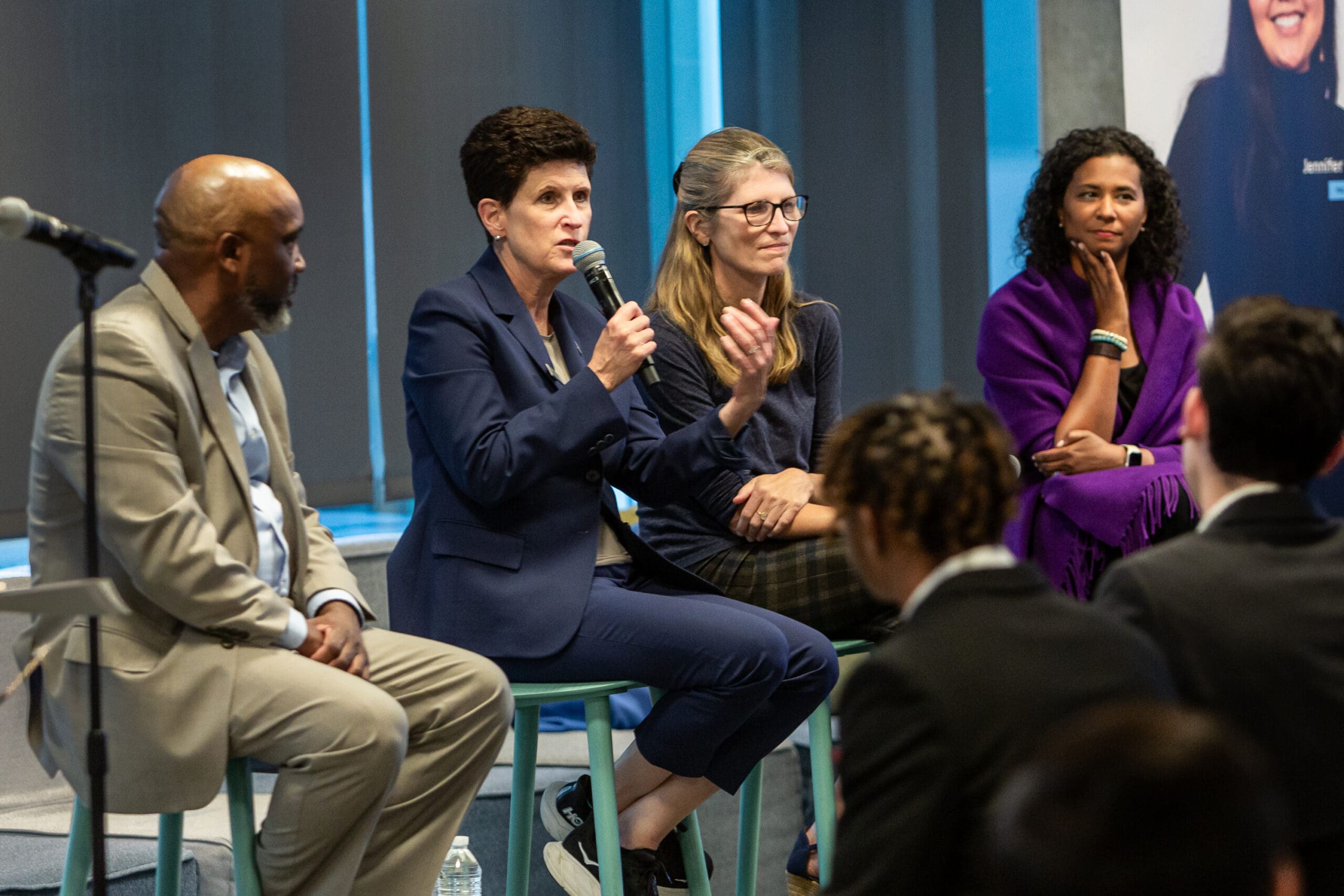 Four individuals on stage participate in a panel discussion, with one person speaking into a microphone. An audience is visible in the foreground.