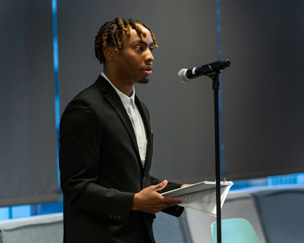 A person in a black suit speaks into a microphone while holding notes. They stand in front of a grey backdrop.