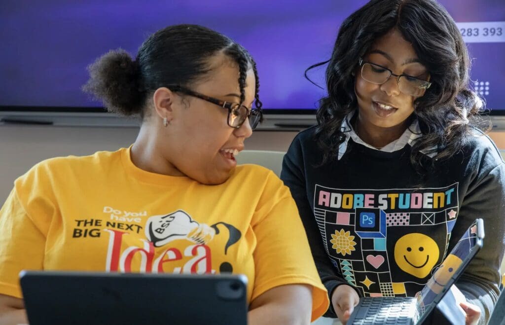 Two women with glasses are looking at a laptop. One wears a yellow shirt with text, and the other wears a black shirt with colorful graphics. Tablets are visible in front of them.