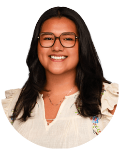 A woman with long dark hair and glasses is smiling at the camera, wearing a white blouse with floral embroidery on the shoulders.