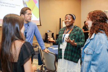 Four people are engaged in conversation in a conference room. A screen displays colorful shapes in the background.