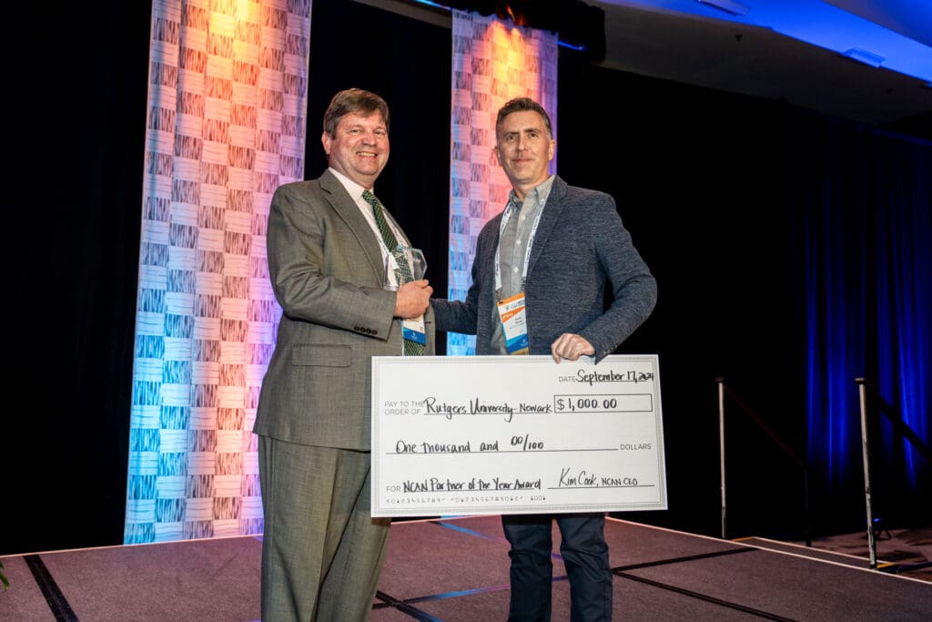 Two men standing on a stage, holding a large ceremonial check for $1,500 made out to "Rutgers University-Newark." They are dressed in formal attire with conference name tags.