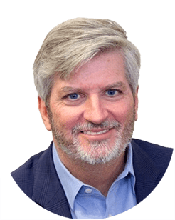 Man with gray hair and beard in a blue shirt and dark jacket, smiling at the camera.