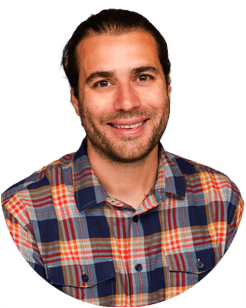 A man with dark hair pulled back, a trimmed beard, and a plaid shirt, smiling at the camera against a plain white background.