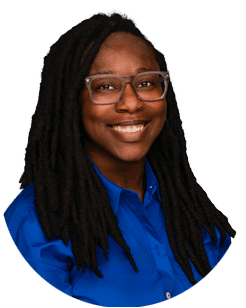 Person with long dreadlocks, glasses, and a blue collared shirt smiling at the camera against a plain background.