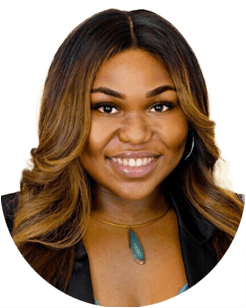 A woman with long, wavy brown hair smiles, wearing a necklace with a blue pendant and a black blazer.