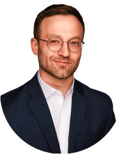Man wearing glasses, a dark suit jacket, and a white shirt, posing against a plain light background.