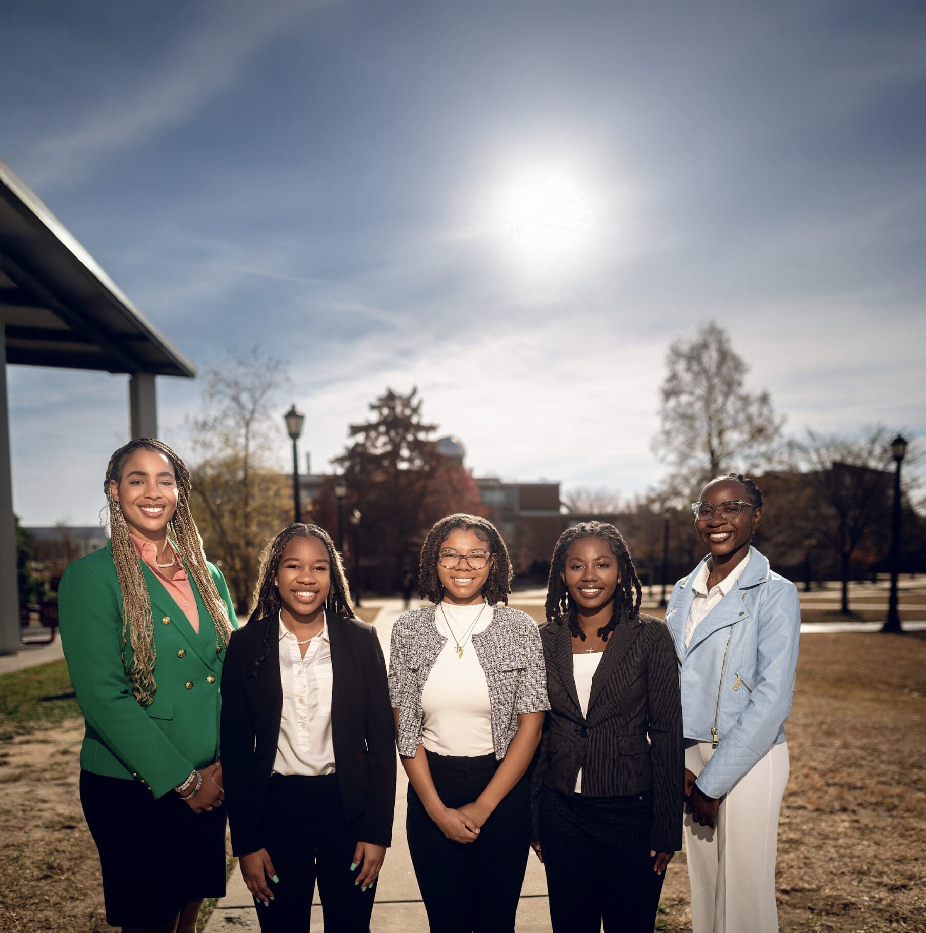 Five people dressed in business attire stand outdoors in a park-like setting under a bright sky, posing for a group photo.