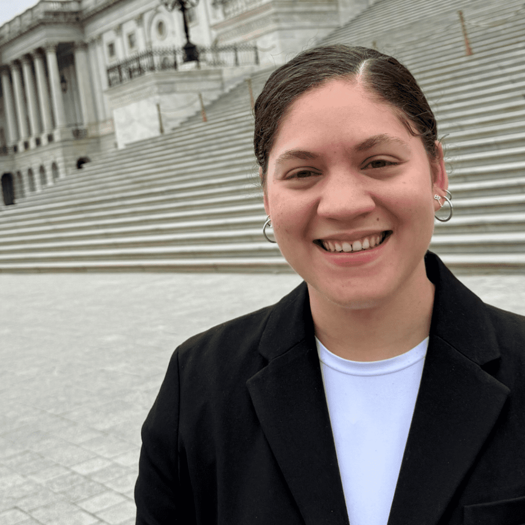 A person wearing a black blazer and white shirt stands smiling in front of large stone steps and a government building.