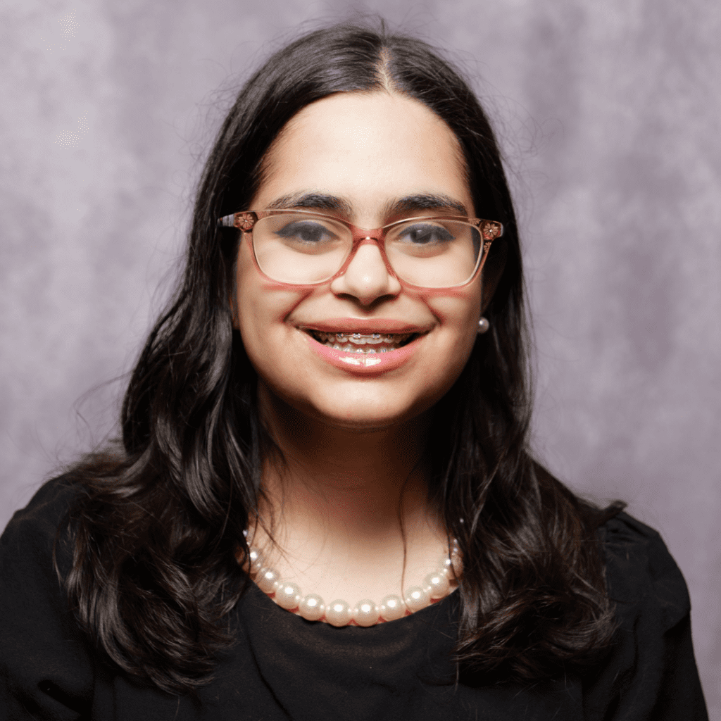 Young woman with long dark hair, wearing glasses, pearl necklace, and black top, smiling in front of a light purple background.