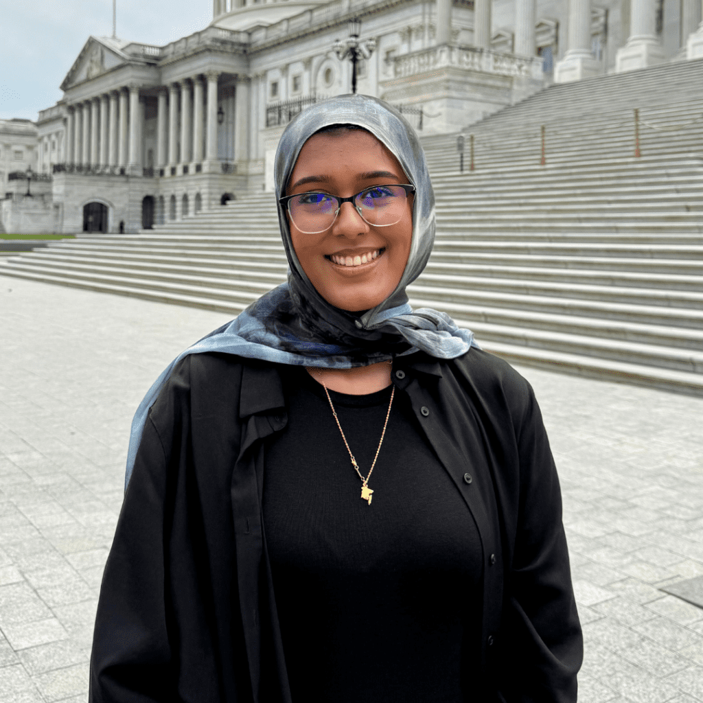 A person wearing glasses and a headscarf stands smiling in front of a large stone building with steps and columns.