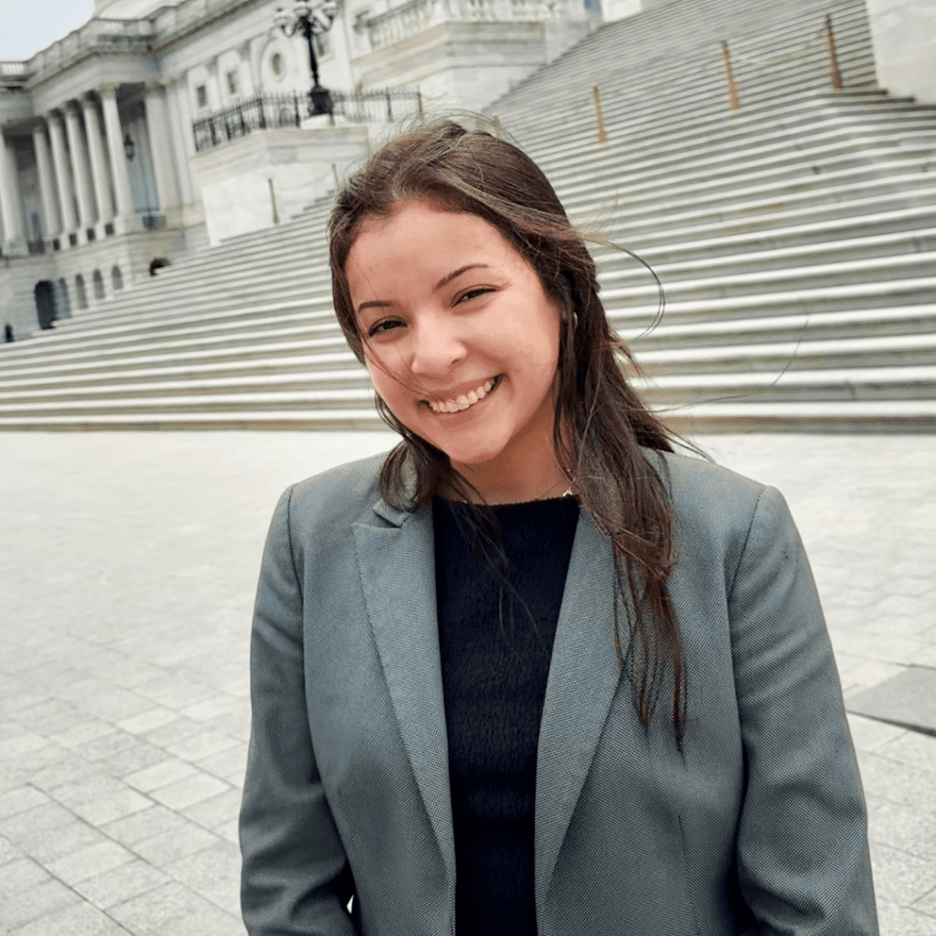 A woman in a gray blazer and black top smiles while standing in front of wide stone steps and a classical government building.