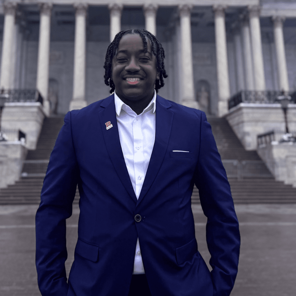 A man in a blue suit stands smiling in front of a large building with columns and wide steps.