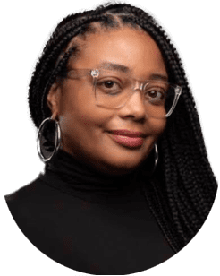 A woman with long braided hair wearing clear glasses, large hoop earrings, and a black turtleneck, smiling at the camera.