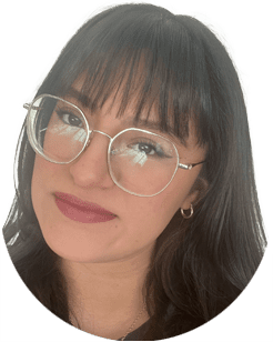 A person with straight dark hair, wearing round eyeglasses and small hoop earrings, is looking at the camera and smiling slightly.