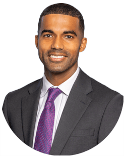 A man in a dark suit, white shirt, and purple tie smiles at the camera against a plain light background.