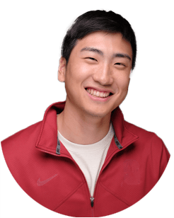A young man with short black hair smiles, wearing a red zip-up jacket over a white t-shirt, against a plain light background.