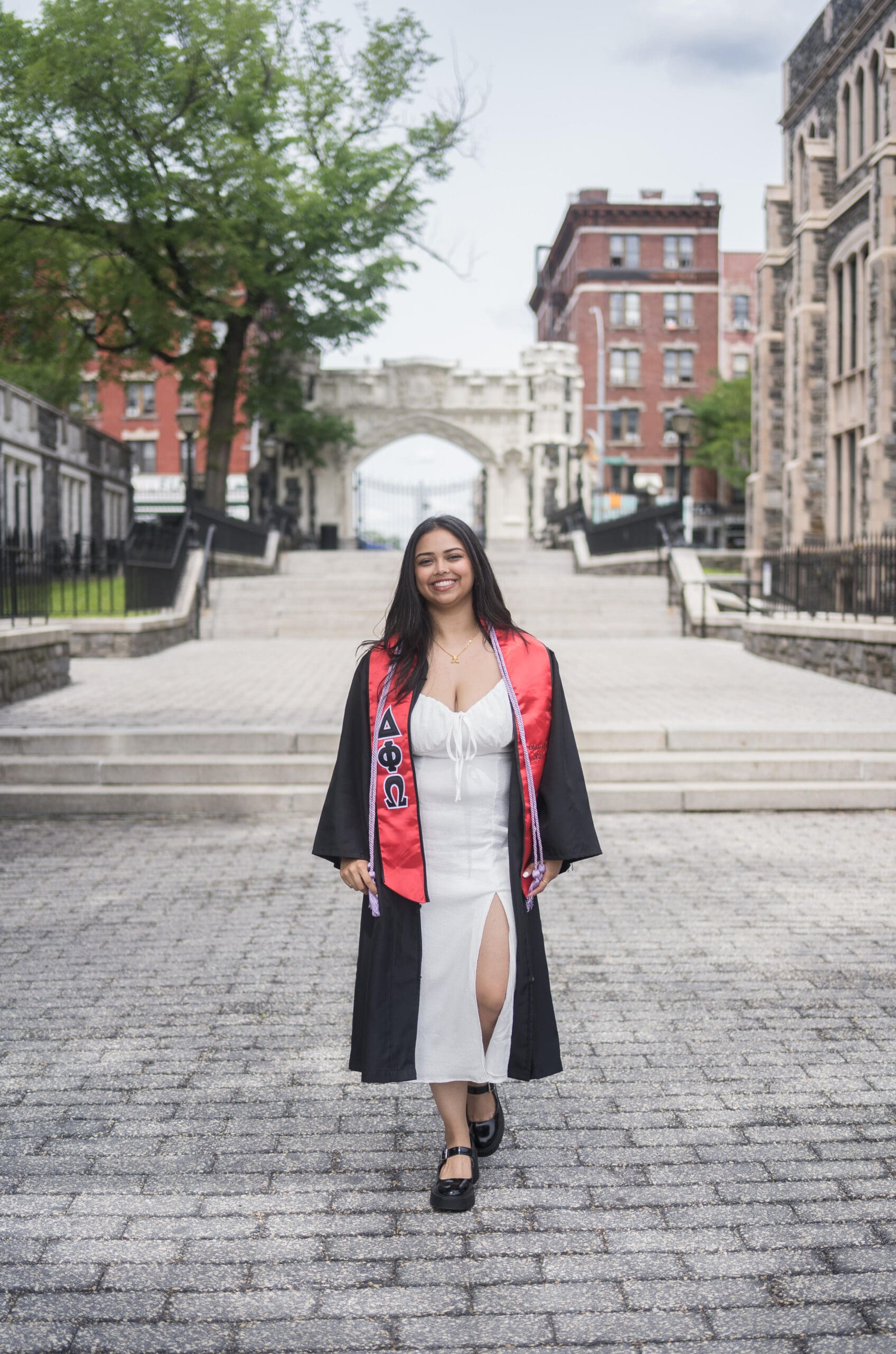 A young woman in a graduation gown and sash stands smiling on a paved walkway with historic buildings and trees in the background.