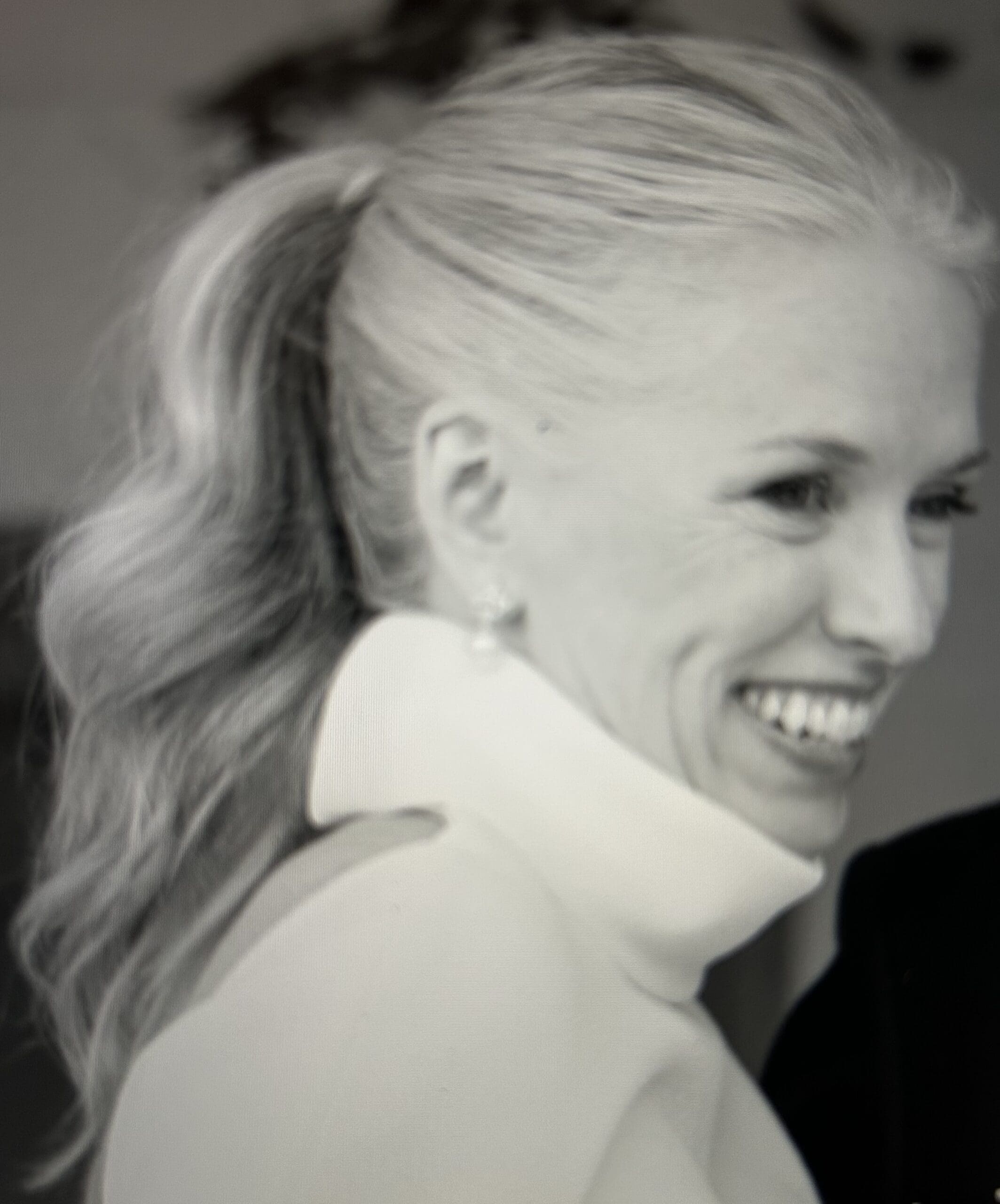 A woman with long, wavy hair in a ponytail smiles, wearing a high-collared garment. The photo is in black and white.