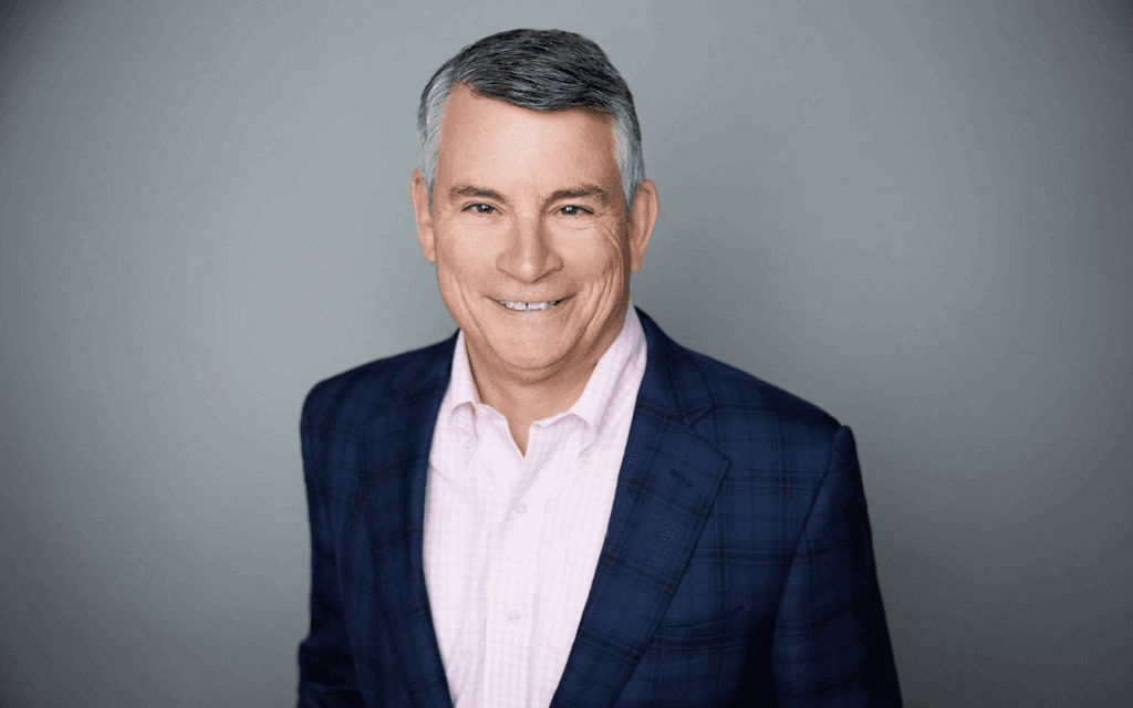 A middle-aged man with short gray hair wearing a navy blue plaid blazer and light pink shirt, smiling in front of a plain gray background.