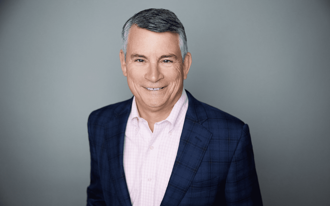A middle-aged man with short gray hair wearing a navy blue plaid blazer and light pink shirt, smiling in front of a plain gray background.