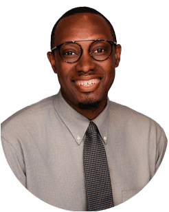 A man wearing glasses, a light gray button-up shirt, and a patterned tie, smiling at the camera against a plain background.