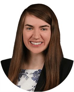 A woman with long straight brown hair, wearing a black blazer over a white blouse with blue floral prints, smiling at the camera against a plain background.