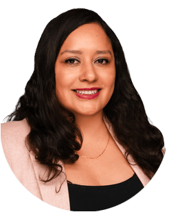 Woman with long dark wavy hair wearing a light pink blazer and black top, smiling at the camera against a plain background.